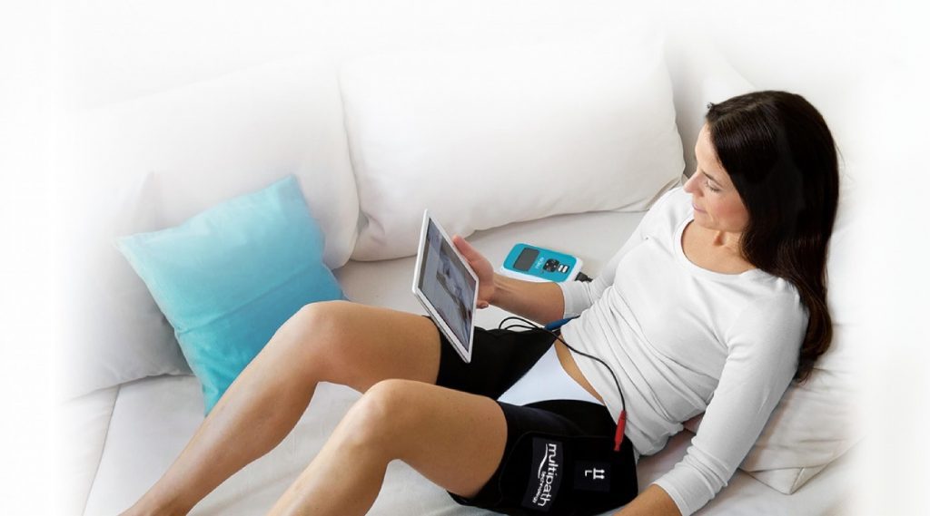 A woman relaxing on a white couch using a tablet, with a blue cushion behind her and a medical device attached to her legs.